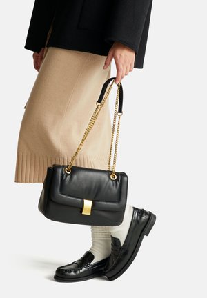 Zwarte leren handtas met een gouden sluiting en kettingband, met een gestructureerde vorm. Draagt met een beige geribbelde rok en zwarte loafers.