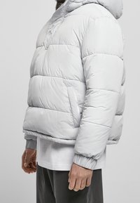 Lysegrå dunjakke med hætte, med quilted design, elastiske manchetter og en frontlomme. Blød, glat tekstur med minimale sømme.
