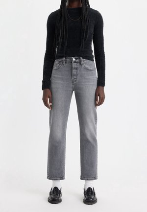 Person iført sort, blød langærmet top, grå straight-leg jeans med høj talje, hvide sokker og sorte læder loafers på hvid baggrund.