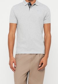 Homme portant un polo à manches courtes gris clair avec un col intérieur à carreaux et un pantalon beige, debout les bras détendus.
