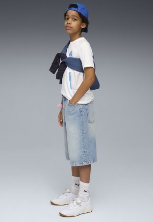 T-shirt bianco con dettagli blu e tasca, maglione blu navy annodato in vita, pantaloni corti in denim azzurro e sneakers bianche.