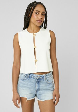 Femme aux cheveux tressés portant un haut blanc sans manches à trois boutons en forme de coquillage et un short en jean bleu clair sur un fond gris.