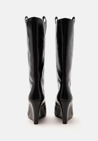 Paire de bottes hautes en cuir noir avec des talons hauts incurvés, photographiées de dos sur fond blanc.