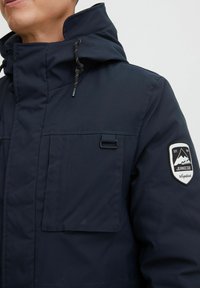 Parka bleu marine avec col montant, capuche avec cordon de serrage réglable et deux poches avant. Présente un patch logo sur la manche.