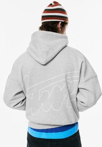 Grijze capuchontrui met een groot wit omlijnd logo op de achterkant. Gecombineerd met een blauw shirt dat eronder gedragen wordt.