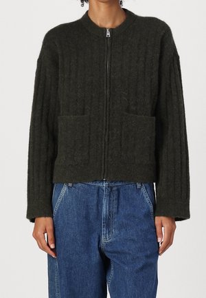 Personne portant un cardigan en maille verte foncée zippée avec poches avant et un jean bleu taille haute sur un fond uni.