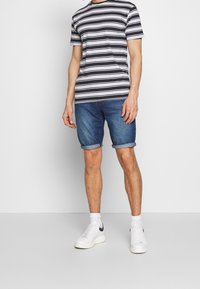 T-shirt a righe con maniche corte bianco e nero, abbinato a pantaloni shorts in denim blu con orlo arrotolato. Sneakers bianche completano l'outfit.
