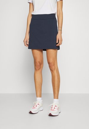 Persoon die een wit t-shirt draagt, een marineblauwe korte rok, witte sokken en witte sneakers met roze accenten, staand tegen een effen achtergrond.