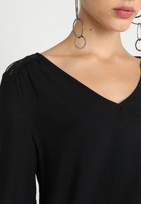 Haut noir avec un col en V, tissu doux et détail en dentelle sur l'épaule. Accessoires: grandes boucles d'oreilles circulaires.