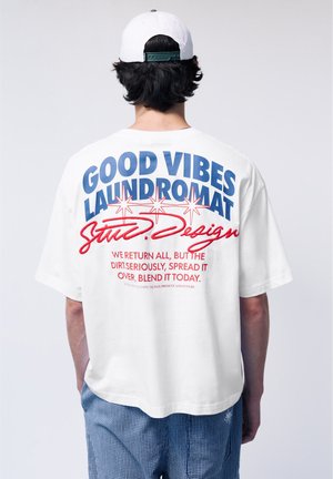 Personne portant une casquette blanche et un t-shirt blanc oversize avec un texte bleu et rouge : « Good Vibes Laundromat » avec un texte supplémentaire.