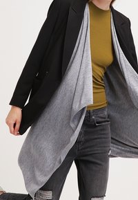 Blazer negro superpuesto a un cárdigan drapeado gris, combinado con una blusa mostaza y jeans negros desgastados. Texturas suaves, ajuste relajado.