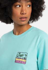 Turquoise cotton t-shirt featuring a square graphic with palm trees, sand, and water in vibrant colors. Classic crew neck design.