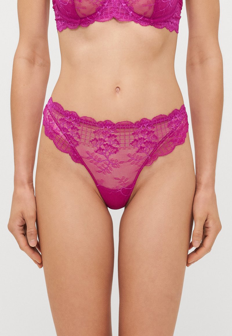 String en dentelle fuchsia avec des motifs floraux, des bords festonnés et un tissu semi-transparent. La ceinture est ornée de détails délicats.