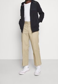 Sudadera con capucha azul marino con cremallera y logotipo, camiseta blanca, pantalones anchos caqui y zapatillas blancas de caña alta, sobre un fondo neutro.