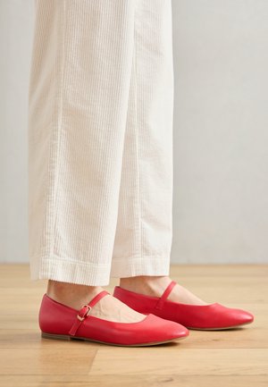 Personne portant un pantalon blanc à jambes larges et des chaussures plates rouges type Mary Jane, debout sur un sol en bois.