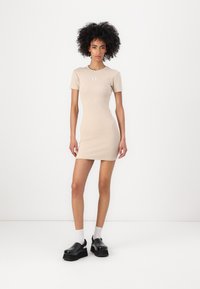 Beige ribbed short-sleeve dress with fitted shape, featuring a small logo patch on the chest. Paired with black chunky platform shoes and white socks.