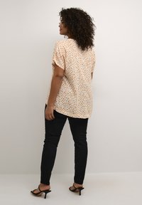 Blouse à manches courtes couleur crème clair avec un motif abstrait orange et marron, associée à un jean noir slim et des sandales noires à talons.