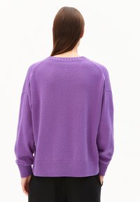 Person mit langen braunen Haaren, die einen lässigen lila Strickpullover und schwarze Hosen trägt, steht mit dem Rücken zu einem schlichten weißen Hintergrund.