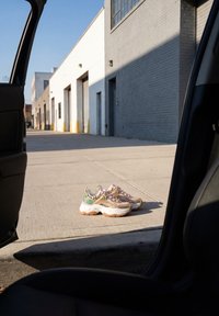 Paire de baskets beige et verte épaisses posées sur un trottoir ensoleillé à l’extérieur d’un bâtiment industriel, vue depuis une porte de voiture ouverte.