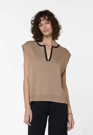 Vrouw met krullend haar, gekleed in een mouwloos beige topje met zwarte afwerking en een zwarte broek, staand tegen een effen witte achtergrond.