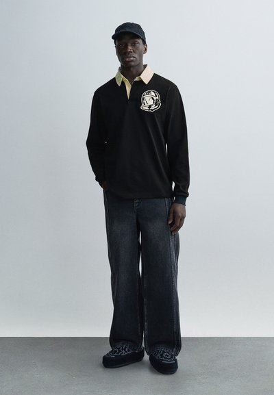 Μαύρο πόλο με μακριά μανίκια και κίτρινο γιακά, με σχέδιο θεματικό για το διάστημα, συνδυασμένο με φαρδιά σκούρα τζιν και μαύρα παπούτσια τύπου slip-on με σχέδια.