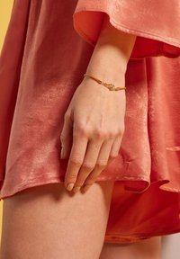 La mano de una mujer descansando cerca del muslo, luciendo una pulsera de oro con una pequeña joya, vestida con una falda o vestido de terciopelo color coral.