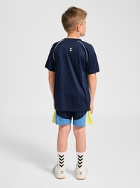 Junge steht mit dem Rücken zur Kamera, trägt ein dunkelblaues Sportshirt, blaue und gelbe Shorts, weiße Socken mit schwarzen Chevron-Mustern und weiße Sneakers.