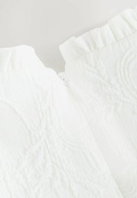 Tessuto bianco texturizzato con motivo floreale e bordi arricciati, con una piccola cerniera bianca parzialmente visibile al centro.