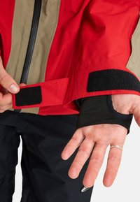 Persoon die een zwart klittenbandsluiting op de mouw van een rood en beige jack aanpast, met een hand die een zilveren ring draagt en zwarte broek.