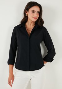Black button-up shirt with a collar, long sleeves rolled at the cuffs, front pockets, and a smooth texture. Paired with white pants.