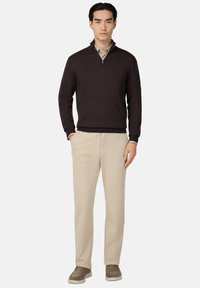 Brūns ar rāvējslēdzēju pulovers, bēšas bikses un pelēkas kurpes. Puloverim ir augsts apkakle; bikses ir taisnas un gludas struktūras.