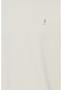 Camiseta de color beige claro hecha de algodón. Presenta un diseño simple con un pequeño símbolo dorado en el área del pecho. Textura suave.
