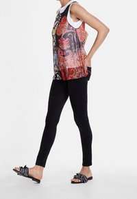 Žena oblečená v mnohobarevném paisley topu bez rukávů, černých úzkých kalhotách a černých sandálech s otevřenou špičkou a bílými popruhy, stojící s rukama v kapsách.