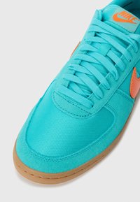 Zapatillas en color turquesa con parte superior texturizada, acentos de ante, logo naranja y cordones planos. Suela de goma marrón claro con detalles de costura.