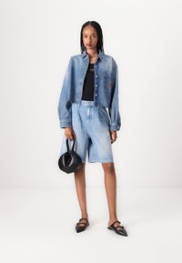 Giacca di denim con design cropped, colore azzurro chiaro e bottoni grandi. Abbinata a pantaloni shorts in denim azzurro chiaro e scarpe flat nere. Borsa nera.
