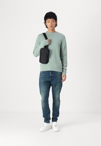 Suéter de malha verde menta, jeans de ganga azuis e ténis brancos. Uma bolsa cruzada preta pende do ombro, contra um fundo simples.
