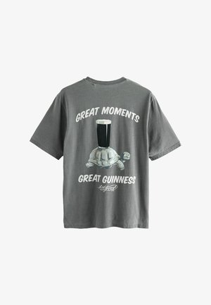 Pilkas medvilnės marškinėliai su vėžlio iliustracija, ant kurios nugaros yra bokalas tamsaus alaus, tekstas sako "Nuostabūs momentai, nuostabus Guinness".