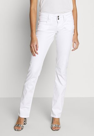 Kobieta w białych, dopasowanych dżinsach z podwójnym zapięciem na guziki oraz sandałach na obcasie z motywem zebry, stojąca na jednolitym tle.