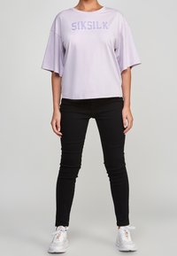 Lila oversized t-shirt med "SIKSILK"-logotyp i en texturerad stil, kombinerad med svarta tighta byxor och vita sneakers. Slät tygkvalitet, avslappnad passform.