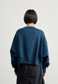 Sötétkék, rövidített kapucnis pulóver, oversized ujjaival, bordázott kerek nyakával és sima textúrával, fekete szoknyával párosítva.