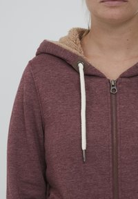 Bordo dukserica s patentnim zatvaračem i bež pamučnom oblogom, sa podešavajućom špagom sa srebrnim vrhovima i zatvaranjem na patent. Teksturirani materijal.
