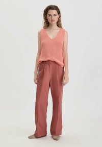 Pfirsichfarbene, texturierte ärmellose Bluse mit V-Ausschnitt, kombiniert mit hochgeschnittenen, weit geschnittenen, rostfarbenen Hosen. Neutrale Schuhe runden den Look ab.