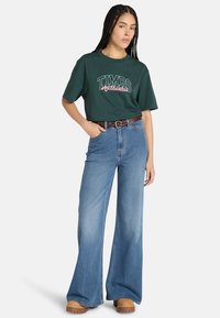Grünes Baumwoll-T-Shirt mit "TIMBS Authentic" Druck, kombiniert mit hoch taillierten, weiten blauen Jeans und einem braunen Gürtel. Braune Holzschuhe an den Füßen.