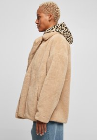 Béžový kabát z umelého kožušiny s kapucňou so leopardím vzorom, s voľným strihom, širokým golierom, bočnými vreckami a jemnou textúrou.