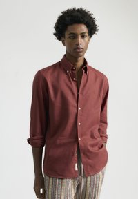 Camisa de botones color burdeos hecha de tejido ligero, con cuello, mangas largas y puños enrollados. Etiqueta destacada en el lateral.
