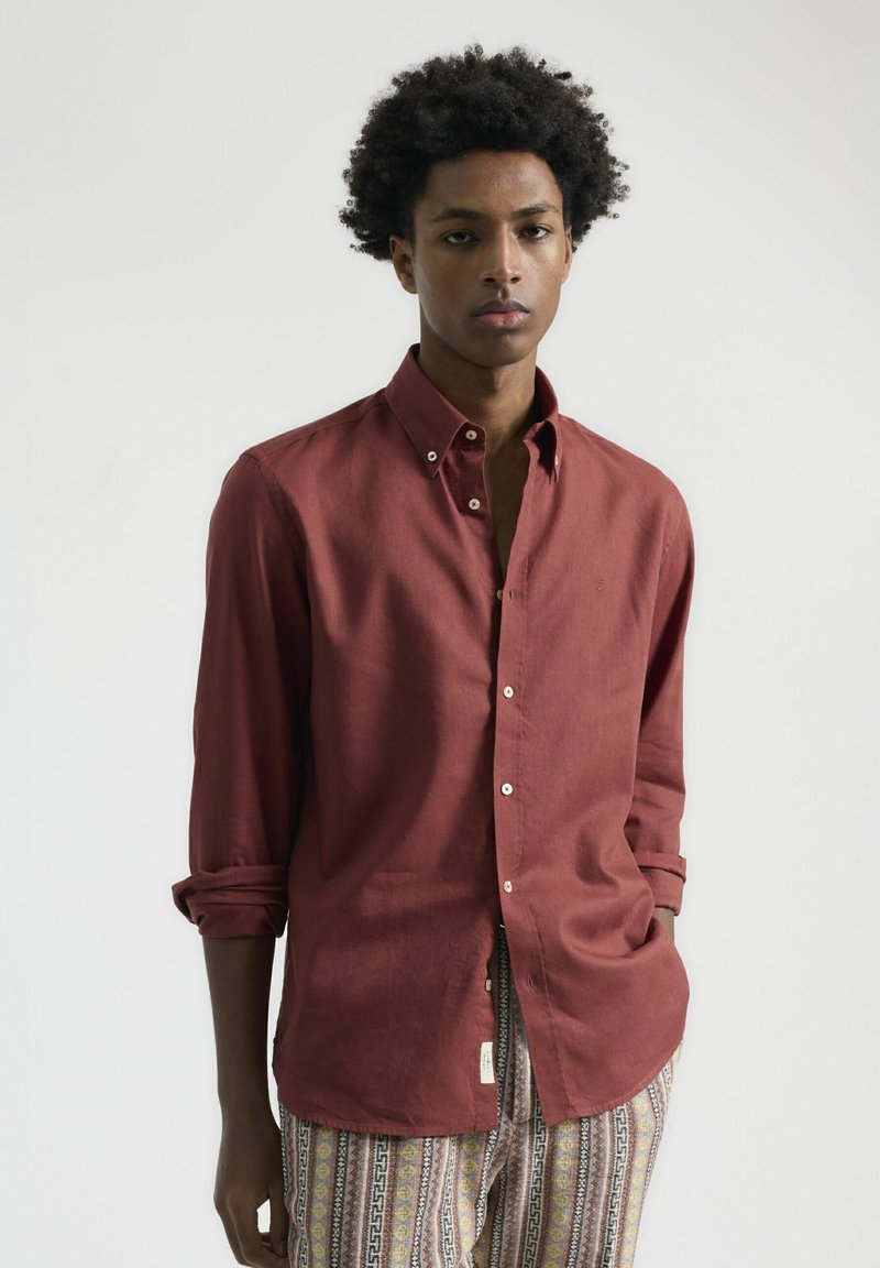 Camisa de botones color burdeos hecha de tejido ligero, con cuello, mangas largas y puños enrollados. Etiqueta destacada en el lateral.