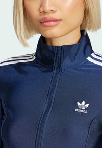 Marinblå zip-up jacka med vitt Adidas-logotyp och tre vertikala vita ränder på axlarna; slätt, glänsande tyg med hög krage.