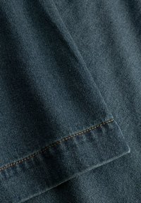 Tmavomodrá rifľovina s jemnou textúrou, s kontrastným oranžovým prešitím pozdĺž lemu, ktoré zvýrazňuje okraj odevu.