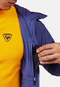 Lila Jacke mit Kapuze und Reißverschlusstasche, getragen über einem strukturierten gelben Hemd mit einem schwarzen Logo. Ein Smartphone wird in die Tasche gesteckt.