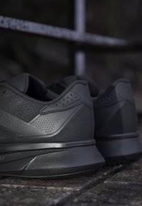 Paire de chaussures de sport gris foncé posées sur une surface en pierre texturée avec une clôture métallique floue en arrière-plan.